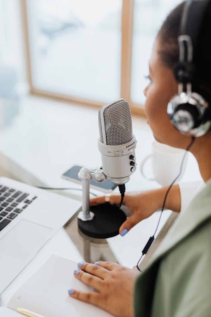 A woman wearing headphones recording a podcast at home using a professional microphone and laptop.
